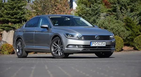 Szary Volkswagen Passat sedan zaparkowany na podjeździe, otoczony zielenią i kamieniami.