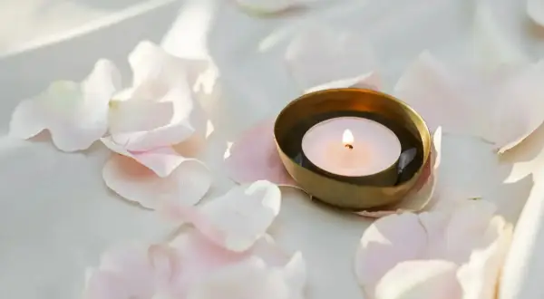 Delikatny płomień tealight pali się w złotej miseczce, otoczony przez jasnoróżowe płatki róż.