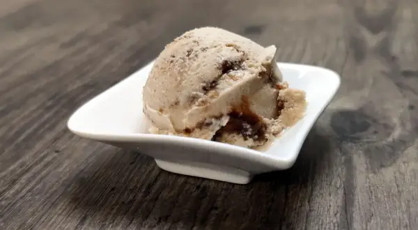 Ein Löffel cremiges Tiramisu Eis mit dunklen Schokoladenstreifen in einer weißen Schale auf Holzhintergrund.