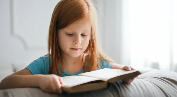 Niña pelirroja concentrada leyendo un libro. Su velocidad lectora por edades es notable para su corta edad.