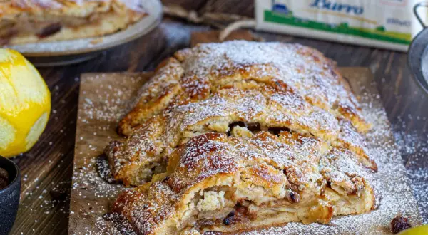 Strudel di pasta frolla dorato, cosparso di zucchero a velo, con ripieno di mele e uvetta. Accanto, scorza di limone e una tazza di uvetta.