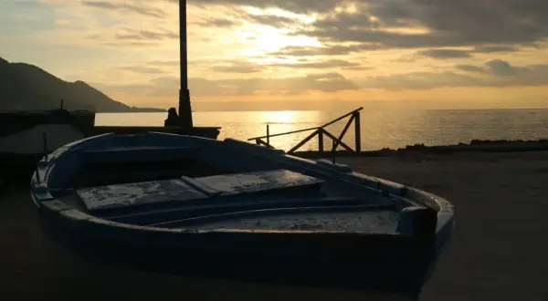 Un'imbarcazione blu riposa sulla riva al tramonto, con il sole che filtra tra le nuvole e illumina il mare calmo.