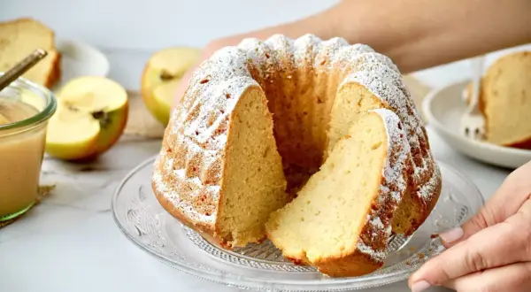 Saftiger Gugelhupf mit Puderzucker bestäubt, daneben Äpfel und ein Glas Apfelmus. Ein Stück Kuchen mit Apfelmus ist ein Genuss.