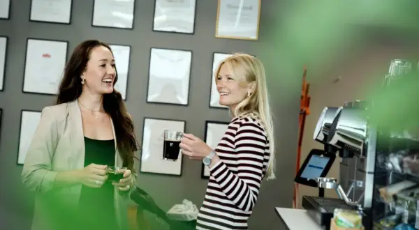 Två kvinnor, en grafisk formgivare, pratar vid en kaffemaskin. Väggen bakom dem är prydd med diplom.
