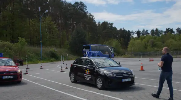 Na placu manewrowym, gdzie obowiązuje ruch lewostronny, czarny samochód szkoleniowy i czerwony pojazd czekają na kursantów. W tle niebieski autokar.