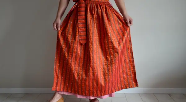 Eine Frau trägt ein Dirndl mit einer leuchtend orangefarbenen Dirndlschürze, die mit Streifen und kleinen Tupfen gemustert ist.