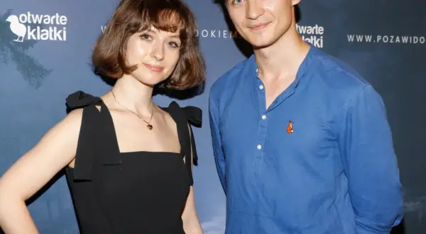 Kasia Sienkiewicz w czarnej sukience i mężczyzna w niebieskiej koszuli z liskiem na piersi pozują na tle niebieskiej ścianki z logo "otwarte klatki".