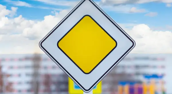 Znak droga z pierwszeństwem, biały romb z żółtym środkiem, na tle błękitnego nieba z chmurami.