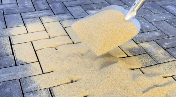 Schaufel mit Sand zum Pflastersteine verfugen. Sand rieselt auf dunkle Steine, um Fugen zu füllen.