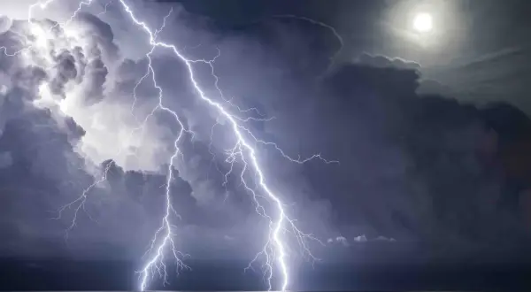 Dramatic lightning photography captures multiple bolts striking the ocean under a stormy sky with a bright moon.