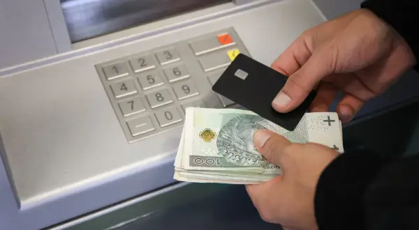 Ręce trzymające banknoty i kartę zbliżają się do wpłatomatu. Pieniądze na koncie czekają na wpłatę.