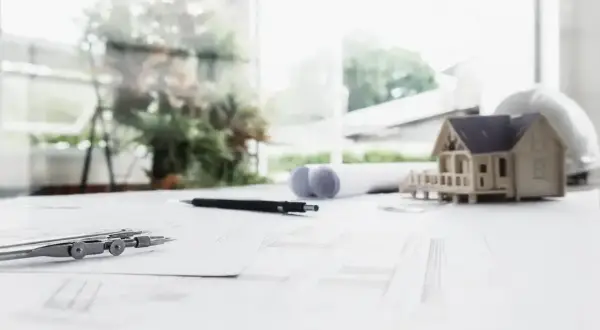 Model domu, cyrkiel, ołówek i plany architektoniczne na biurku. W tle widać zieleń i budynki, sugerujące drugą linię zabudowy.