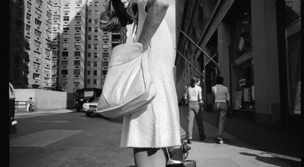 Una donna con borsa a tracolla cammina per strada, con palazzi alti sullo sfondo. Mark Cohen photography cattura la vita urbana.