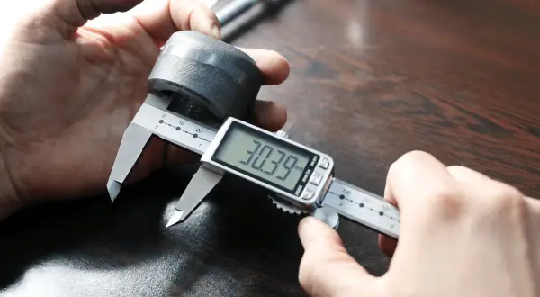 Dłoń trzymająca suwmiarkę cyfrową mierzącą metalowy element.