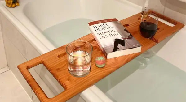 Tabla de madera sobre bañera con libro, vaso de agua, bomba de baño y copa de vino. Preparando un baño relajante.