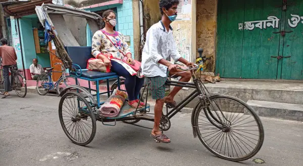 Rowerzysta w masce wiezie kobietę w rikszy. Typowy transport w Indiach, uliczny krajobraz z psami i budynkami.