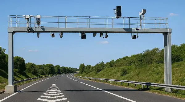Portique de péage sans barrière sur une autoroute bordée d'arbres. Des caméras et des feux sont installés sur la structure métallique.