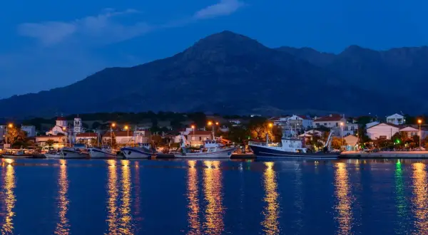 Port z samotraki nocą. Łodzie cumują przy nabrzeżu, a światła odbijają się w spokojnej wodzie. W tle widać góry.