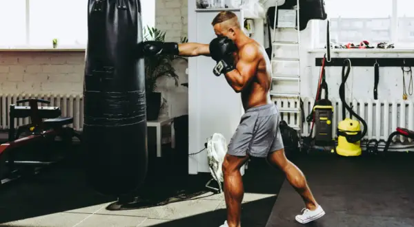 A muscular man punches a heavy bag in a gym, proving that boxing builds muscle.