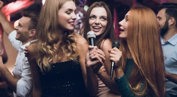 Trzy kobiety śpiewają do mikrofonów, bawiąc się przy karaoke. W tle widać innych ludzi tańczących.