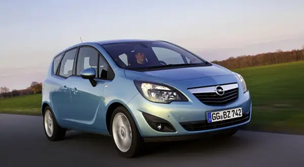 Niebieski Opel Meriva 1.4 benzyna w ruchu, z kierowcą za kierownicą, na tle zielonych pól i drzew.
