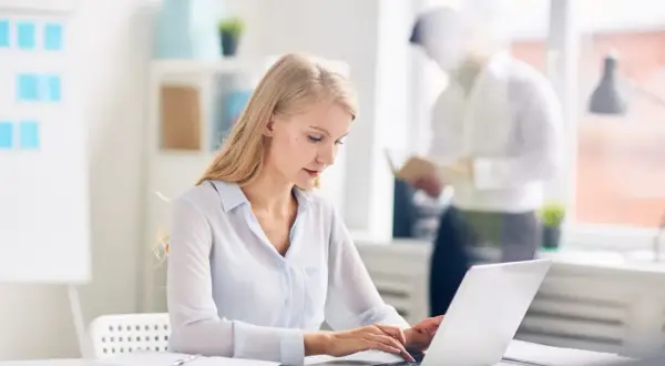 Kobieta pracuje na laptopie w nowoczesnym biurze. W tle mężczyzna czyta dokumenty. Nowoczesne biuro z office 2021.