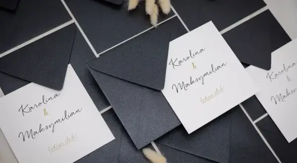 Elegancki zaproszenie wzór na ślub Karoliny i Maksymiliana. Białe karty z czarnym i złotym napisem, otoczone czarnymi kopertami.