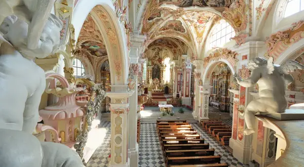 Prunkvolle Deckenmalereien und Stuckaturen im Kloster Neustift. Engelstatue blickt auf Kirchenbänke und den Altar.