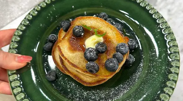 En hög med amerikanska pannkakor toppas med smör, blåbär och sirap på en grön tallrik.