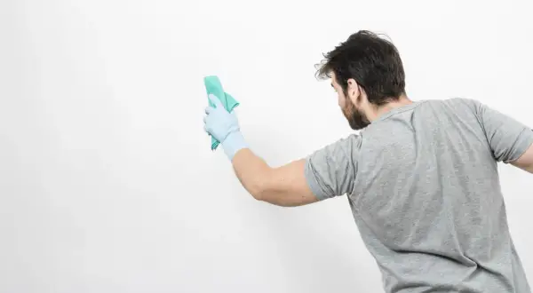 Un homme en gant bleu nettoie un mur blanc avec un chiffon vert. Il montre comment nettoyer un mur blanc sans enlever la peinture.