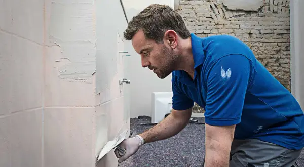 Mann spachtelt Fliesen mit einer Kelle. Er trägt Handschuhe und ein blaues Polohemd.