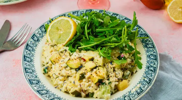 Un crémeux risotto courgette, garni de roquette fraîche et d'une tranche de citron, sur une assiette ornée.