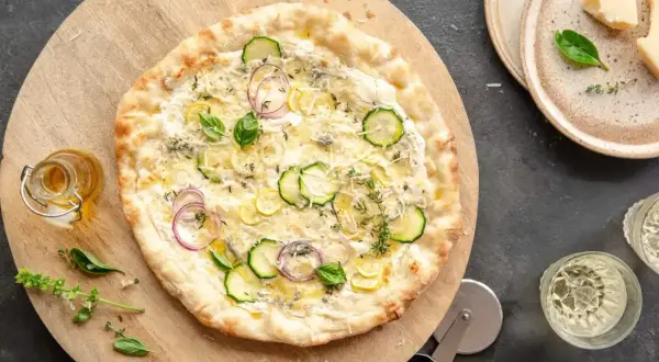 Pizza bianca z cukinią, cebulą i cytryną. Obok buteleczka oliwy i kieliszki wina.