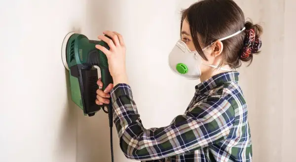Mujer con mascarilla lijando paredes con una lijadora eléctrica.