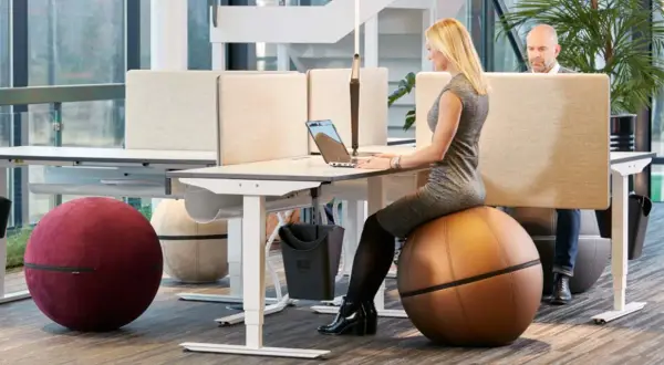Kvinna arbetar vid ett höj- och sänkbart skrivbord i ett aktivitetsbaserat kontor. Hon sitter på en stor, brun träningsboll.