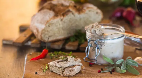 Pasztet na chlebie z ziołami, słoik pasztetu, chleb i papryczka chili na desce.