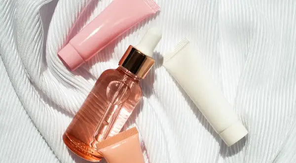 Un frasco de serum con gotero y tubos de crema rosa, blanco y melocotón sobre tela blanca. ¿Que va primero el serum o la crema?