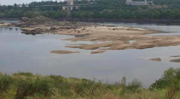 Piaszczysta wyspa z kamieniami na rzece, przez którą płynie. W oddali widoczny most i zabudowania przemysłowe.