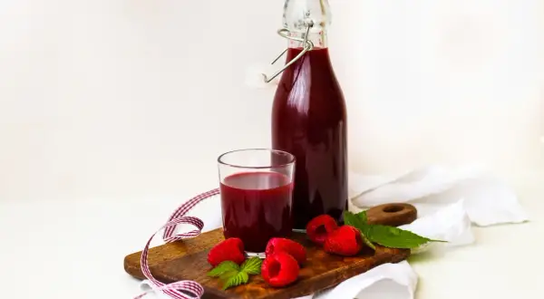 Szklanka i butelka z domowym sokiem z malin, świeże maliny i listki mięty na desce. Idealne na lato, pokazują, jak zrobić sok z malin.