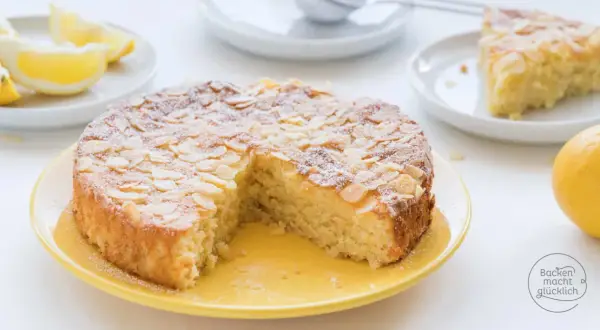 Saftiger Kuchen ohne Mehl, bestreut mit Mandelblättchen und Puderzucker. Dazu Zitronenspalten und eine ganze Zitrone.