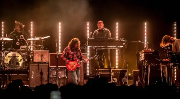 The War on Drugs band performs live, bathed in warm stage lights. A guitarist in a red plaid shirt plays center stage, with a drummer, keyboardist, and another musician on synths.