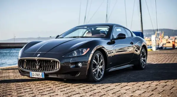 Eleganckie, czarne coupe auto Maserati stoi na nabrzeżu, z żaglami łodzi w tle.