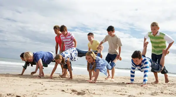Niños compiten en juegos en la playa sin material, haciendo carreras de carretilla humana y otros desafíos divertidos.