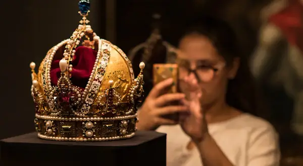 Złota korona, ozdobiona perłami i rubinami, przypomina wielki mogoł diament. Kobieta w okularach robi jej zdjęcie telefonem.