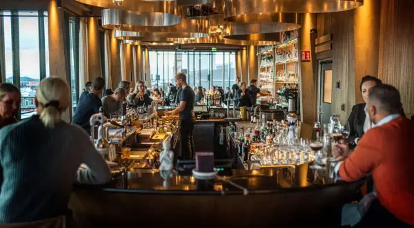 Människor sitter vid en bar med många flaskor och glas. Belysningen är varm och stämningen inbjuder till att läsa **spesso recensioner** om stället.