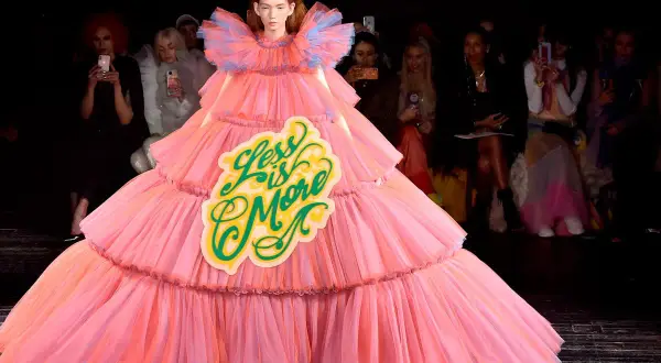 A model wears a voluminous pink tiered gown with a "Less is More" sign, embodying a bold camp aesthetic on the runway.