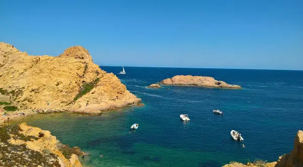 En solig dag vid Ile Rousse. Turkost vatten, klippor och båtar i bukten. En segelbåt syns i fjärran.