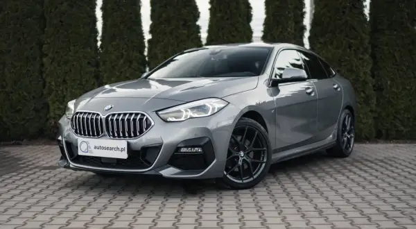 Szary BMW 218i z czarnymi felgami stoi przed żywopłotem.
