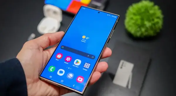 Mano sujetando un smartphone con pantalla azul vibrante, mostrando iconos de aplicaciones y el tiempo. La interfaz, **que es One UI**, se ve fluida y organizada.