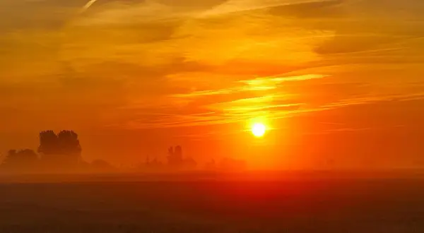Een gloedvolle zonsopgang boven een mistig veld. De zon, een heldere bol, kondigt het wakker worden van de dag aan, met oranje en gele tinten die de hemel vullen.